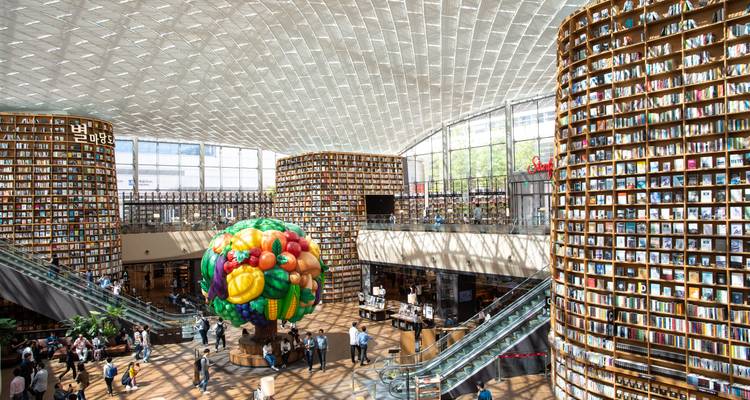 Grande salle de bibliothèque moderne avec des piliers élancés remplis de livres, un toit en verre et une sculpture artistique colorée, animée par de nombreux visiteurs.