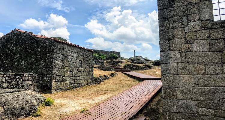 Steinhäuser und ein hölzerner Gehweg schlängeln sich durch ein rustikales Hügeldorf unter einem hellen Himmel
