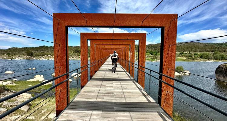 Ein Radfahrer fährt durch sich wiederholende rostrot Stahlrahmen auf einer Brücke, die sich über einen blauen See spannt