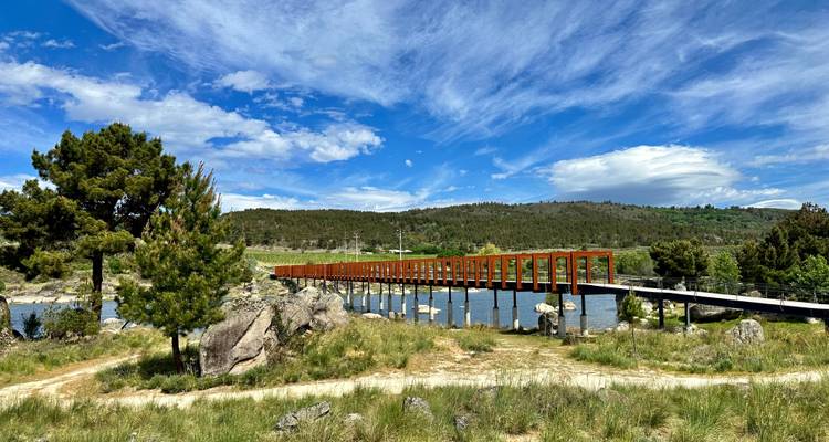 Eine lange moderne Fußgängerbrücke mit rostfarbenen Rahmen überquert einen breiten Fluss inmitten grüner Hügel