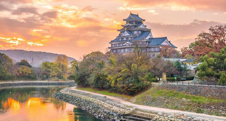El Castillo de Okayama al atardecer dorado con vista a un foso reflectante bordeado de follaje colorido
