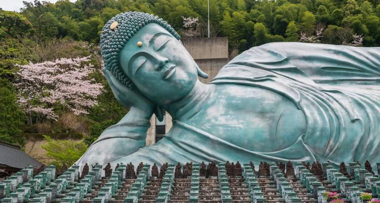 Massive liegende Bronze-Buddha-Statue, die inmitten bewaldeter Hügel und Frühlingsblüten ruht.