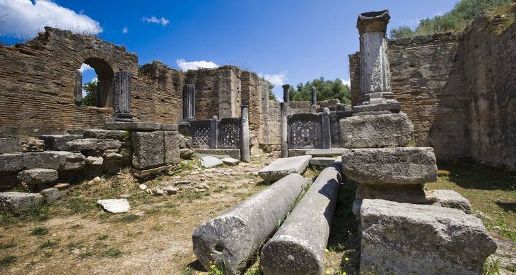 Ruinas de piedra y columnas caídas de la antigua Olimpia bajo un cielo azul despejado.