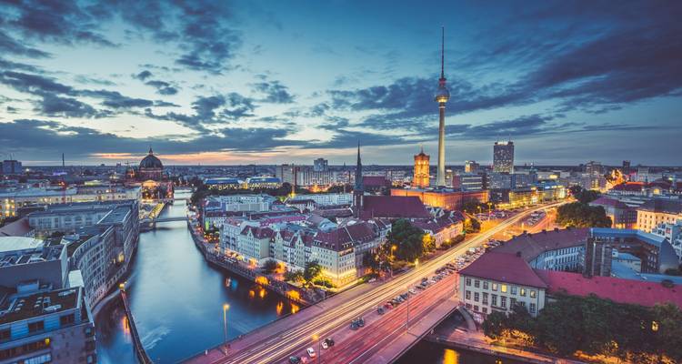 Vista aérea crepuscular sobre un paisaje urbano con la Torre de TV Fernsehturm, calles iluminadas y río serpenteando a través de edificios modernos e históricos