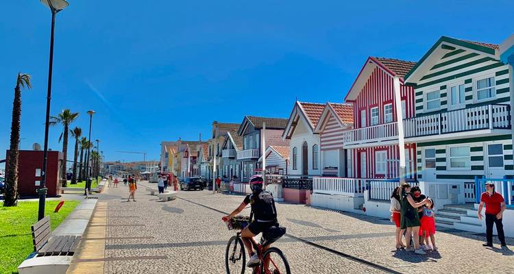 Iconic striped wooden houses of Costa Nova line a sunny pedestrian street with cyclists and strolling families.