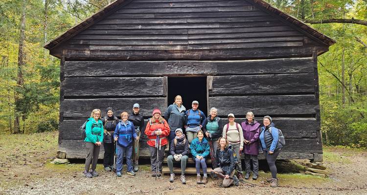 Un grand groupe de randonneurs pose devant une cabane historique en bois sombre dans la forêt.