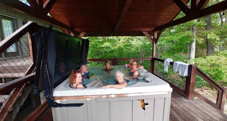 Six femmes se détendent dans un bain à remous extérieur sur une terrasse en bois entourée de verdure estivale.
