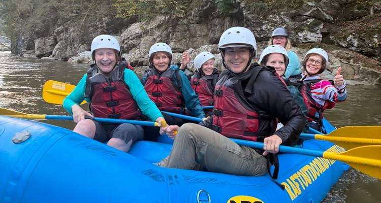 Un groupe joyeux en casques et gilets de sauvetage est assis dans un radeau bleu sur une rivière forestière, prêt pour l'action en eaux vives.