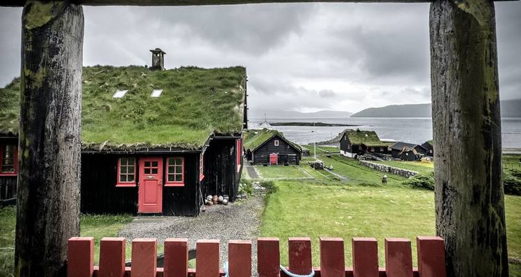 Traditional turf-roof cottages framed by a wooden gate overlook a fjord beneath moody skies.