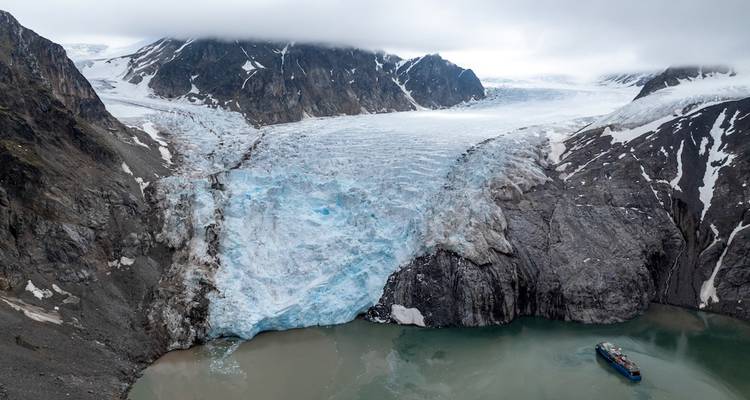 Aerial view of vast blue glacier cascading into a fjord with a small expedition ship below.