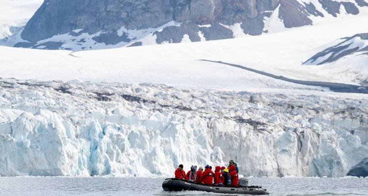 Zodiac full of bundled travelers in red jackets drifts near a towering white glacier wall.