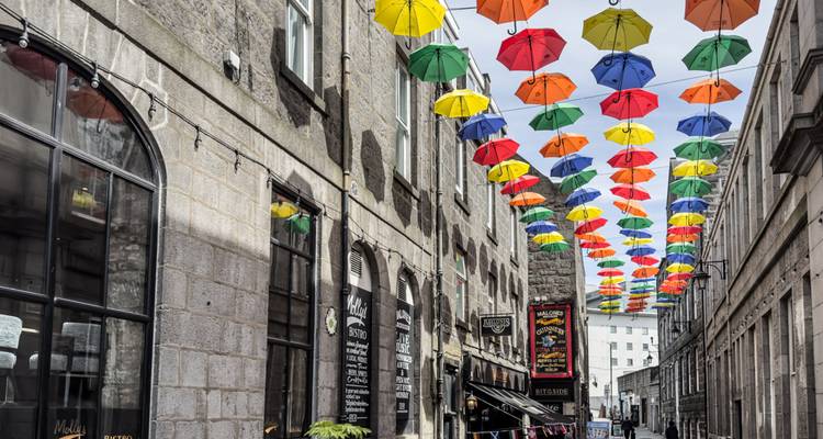 Narrow Aberdeen street decorated with a canopy of colourful umbrellas between granite buildings.