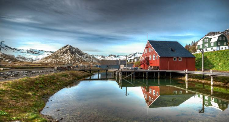Estanque inmóvil reflejando una casa de madera roja y picos cubiertos de nieve bajo un cielo suave y parcialmente nublado.