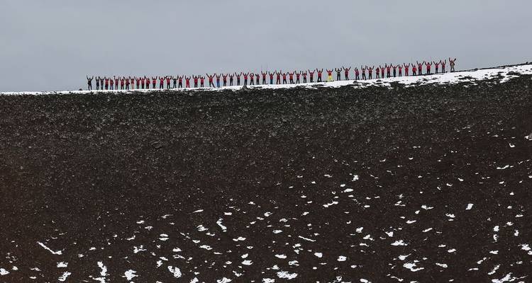 Larga fila de viajeros vestidos con colores brillantes celebrando en el borde de una ladera volcánica árida con nieve irregular.