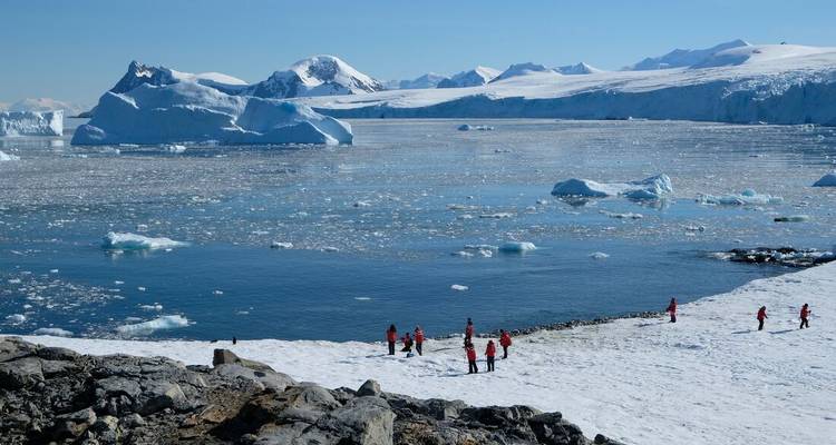Un pequeño grupo con parkas rojas explora una costa antártica helada con icebergs a la deriva en alta mar.