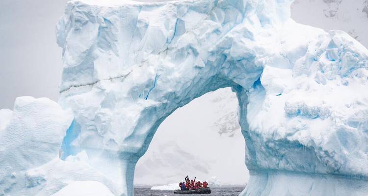Kleines Schlauchboot mit zusammengedrängten Passagieren fährt unter einem hoch aufragenden blauen Eisbogen hindurch.