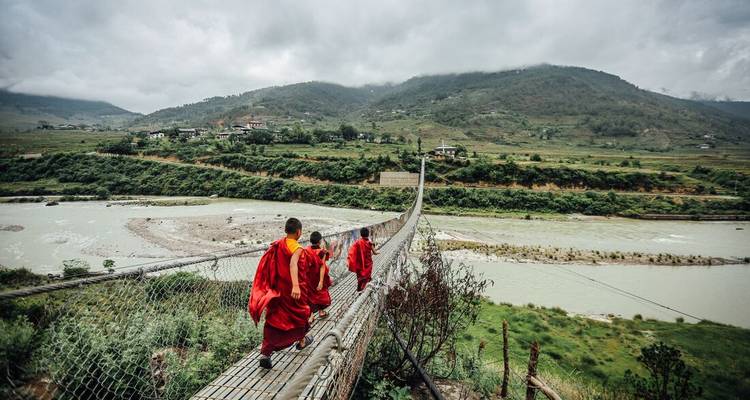 Des moines en robes rouges marchent le long d'un long pont suspendu au-dessus d'une rivière laiteuse dans la luxuriante vallée de Punakha.