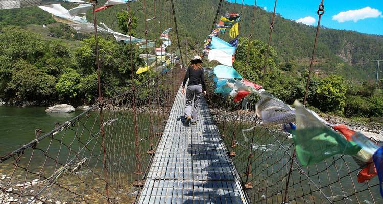Le voyageur traverse un pont suspendu étroit bordé de drapeaux de prière au-dessus des gorges boisées de la rivière.
