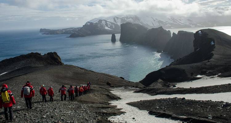 Eine lange Reihe von Entdeckern in roten Parkas schlängelt sich entlang eines schwarzen Vulkanrückens mit Blick auf eine dramatische Caldera und das Meer.