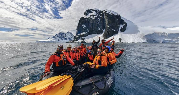 Eine jubelnde Gruppe in orangen Trockenanzügen posiert in einem Schlauchboot mit gelben Paddeln vor hoch aufragenden antarktischen Klippen und sonnenbeschienenen Meeren.