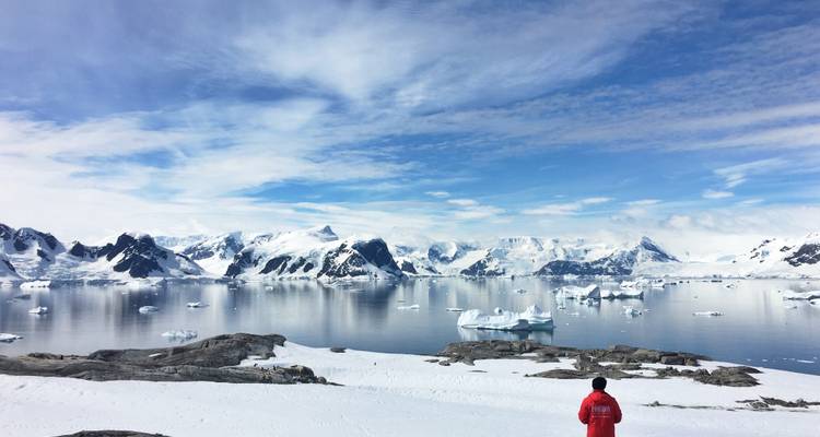 A lone traveler in a red jacket stands on a snowy ridge overlooking mirrored Antarctic waters and dramatic white mountains under a bright sky.