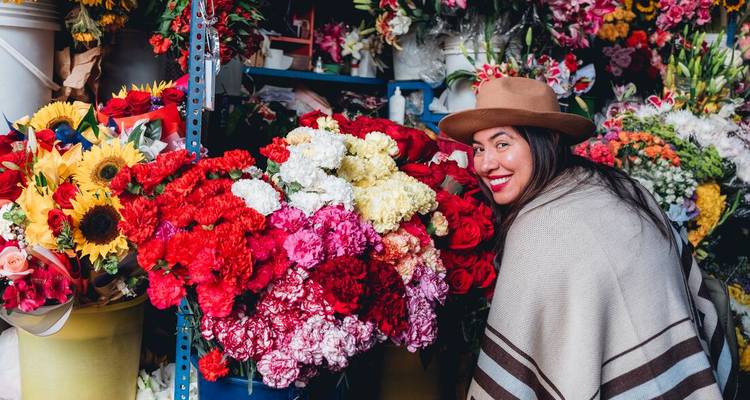A woman in a poncho smiles while surrounded by vibrant buckets of fresh flowers at a local market stall.