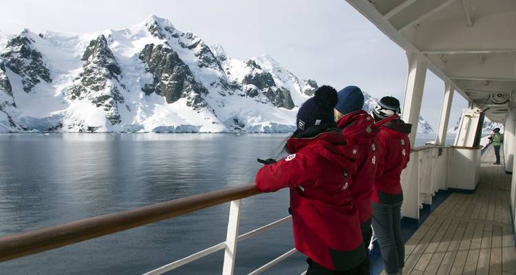 Passagiere in roten Parkas beobachten schneebedeckte antarktische Berge vom Deck eines Expeditionsschiffs.