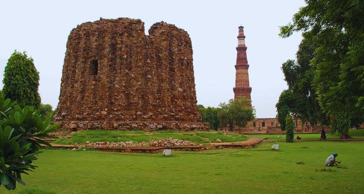 Ruines d'une ancienne tour en pierre avec le majestueux Qutub Minar en arrière-plan au milieu de pelouses luxuriantes.