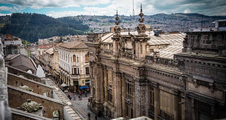 Vista desde la azotea de los edificios históricos ornamentados de Quito y las calles estrechas con gente abajo y colinas al fondo.