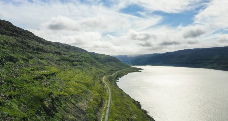 Een kronkelende kustweg loopt langs een lange fjord onder glooiende groene heuvels en wolken die hier en daar verschijnen.
