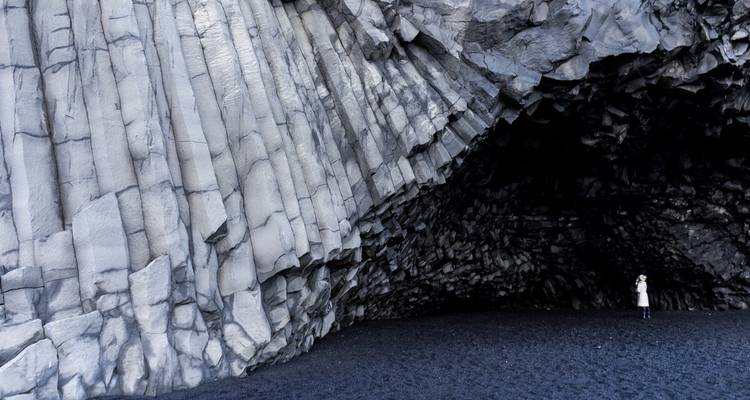 Un visiteur solitaire se tient éclipsé à côté de majestueuses colonnes de basalte gris à l'intérieur d'une grotte sur une plage de sable noir.