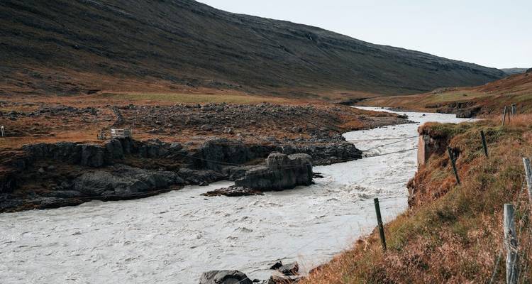 Un fleuve au cours rapide traverse une vallée brune avec un petit pont de bois enjambant les rapides blancs.