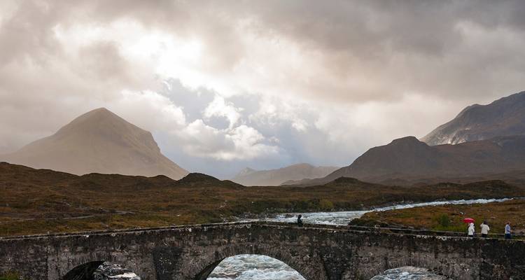 Un vieux pont de pierre enjambe une rivière rapide sous des cieux dramatiques et changeants et des sommets montagneux brumeux.