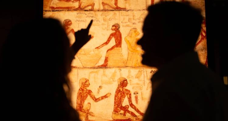 Silhouetted visitors admire ancient Egyptian hieroglyphic art in a softly lit museum gallery.