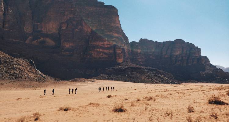 Tiny silhouettes of hikers cross the wide sandy plain beneath towering sandstone cliffs in Wadi Rum.