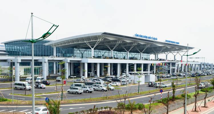 Modern glass-and-steel terminal building of Noi Bai Airport with car park and roadways in foreground on a bright day.