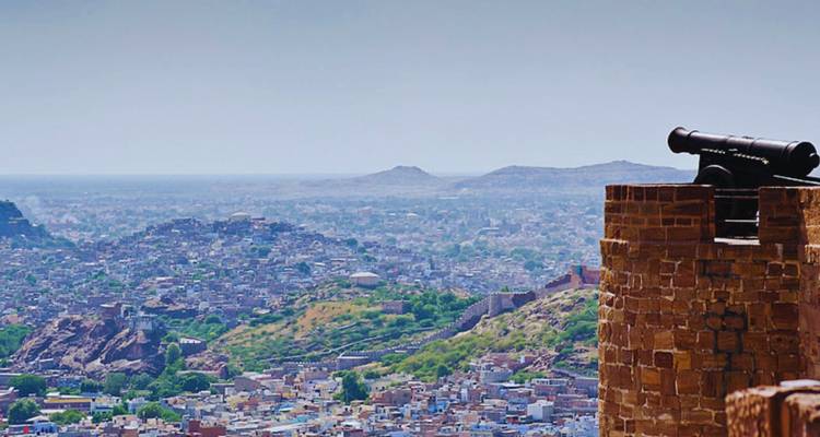 A black cannon atop Mehrangarh Fort towers over the blue cityscape of Jodhpur stretching to distant hills.