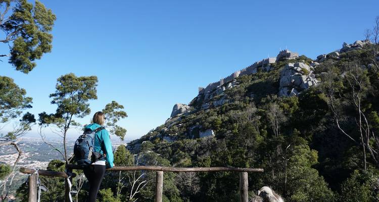 Ein Wanderer macht eine Pause an einem Holzgeländer mit Blick auf die maurische Burg auf einem bewaldeten Hügel.
