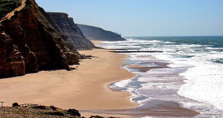 Langer Sandstrand, gesäumt von schroffen Klippen und schäumender Brandung.