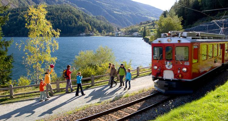 Les randonneurs marchent sur un sentier en bordure du lac tandis qu'un train rouge vintage passe près des eaux alpines cristallines.