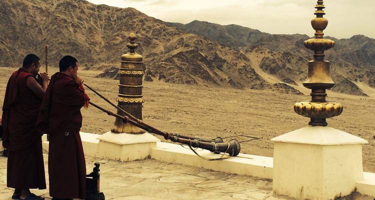 Two monks blow long ceremonial horns from monastery terrace over barren mountain landscape