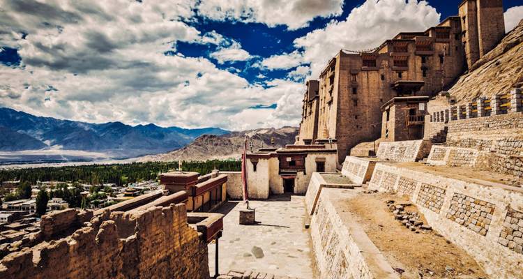 Panoramic view of Leh Palace overlooking the city with dramatic Himalayan mountains under a partly cloudy sky.