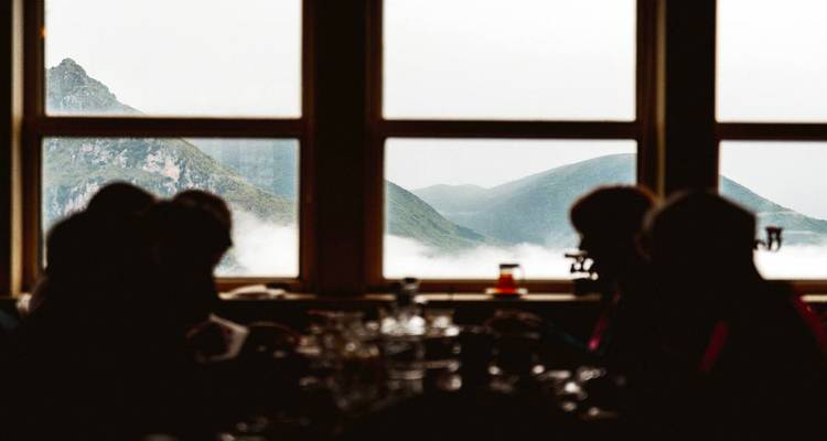 Comensales silueteados dentro de un refugio de montaña contemplan a través de grandes ventanas los picos brumosos.