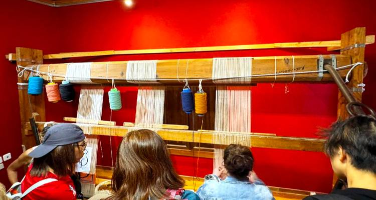 Los visitantes observan una demostración de telar de madera tradicional contra una pared roja vívida.