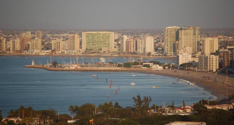 Coastal skyline of high-rise buildings lining a calm bay with small sailboats and sandy shore.