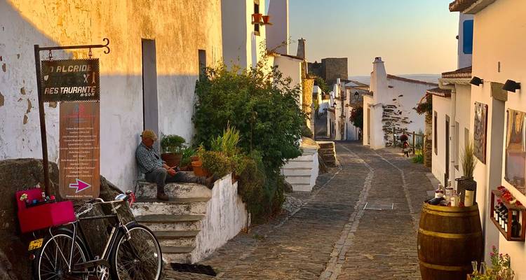 Ein älterer Mann sitzt auf einer Steinstufe neben einem Fahrrad entlang einer charmanten schmalen Dorfstraße in der Abenddämmerung.
