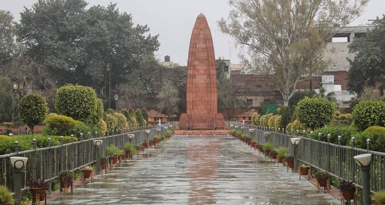 Rain-soaked pathway flanked by manicured shrubs leading to the red sandstone Jallianwala Bagh memorial obelisk.