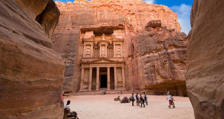 Iconic view of the Treasury at Petra framed by towering red sandstone walls with tourists exploring the forecourt.
