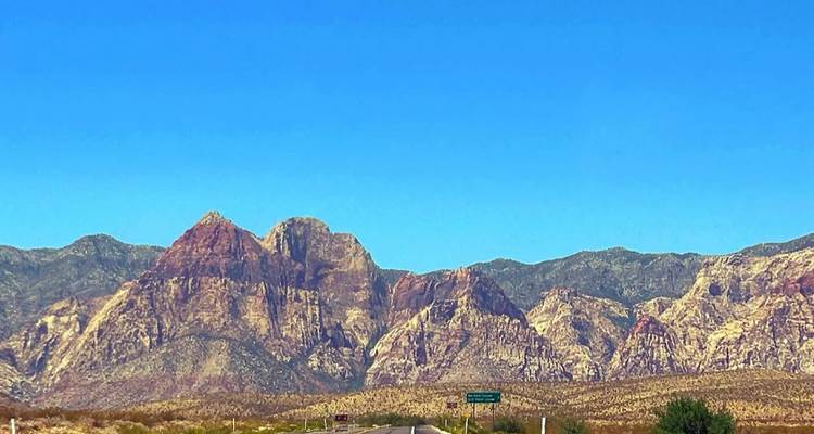 Desert highway leading toward rugged multicoloured mountains beneath a clear blue sky.