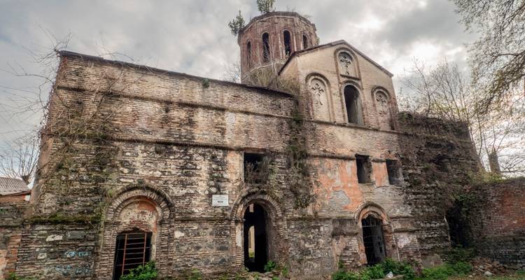Ruine de monastère en pierre usée avec des fenêtres en arc de cercle entourées d'arbres sans feuilles sous un ciel gris.